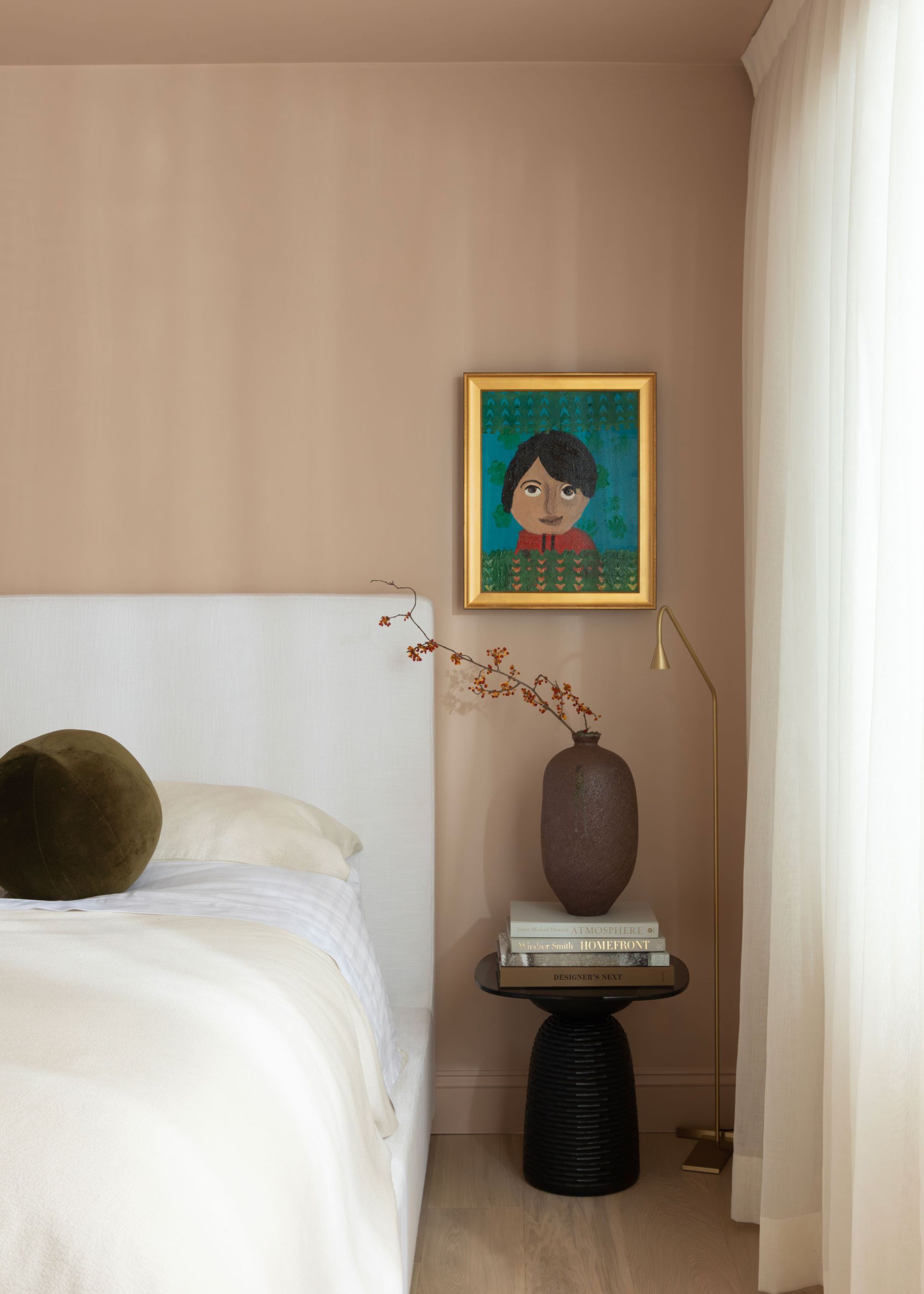 A bedroom painted in dusty pink with a view of the bed with pillows on top. Beside the bed is a bedside table with books and a vase on top. A painting also hands above the table.