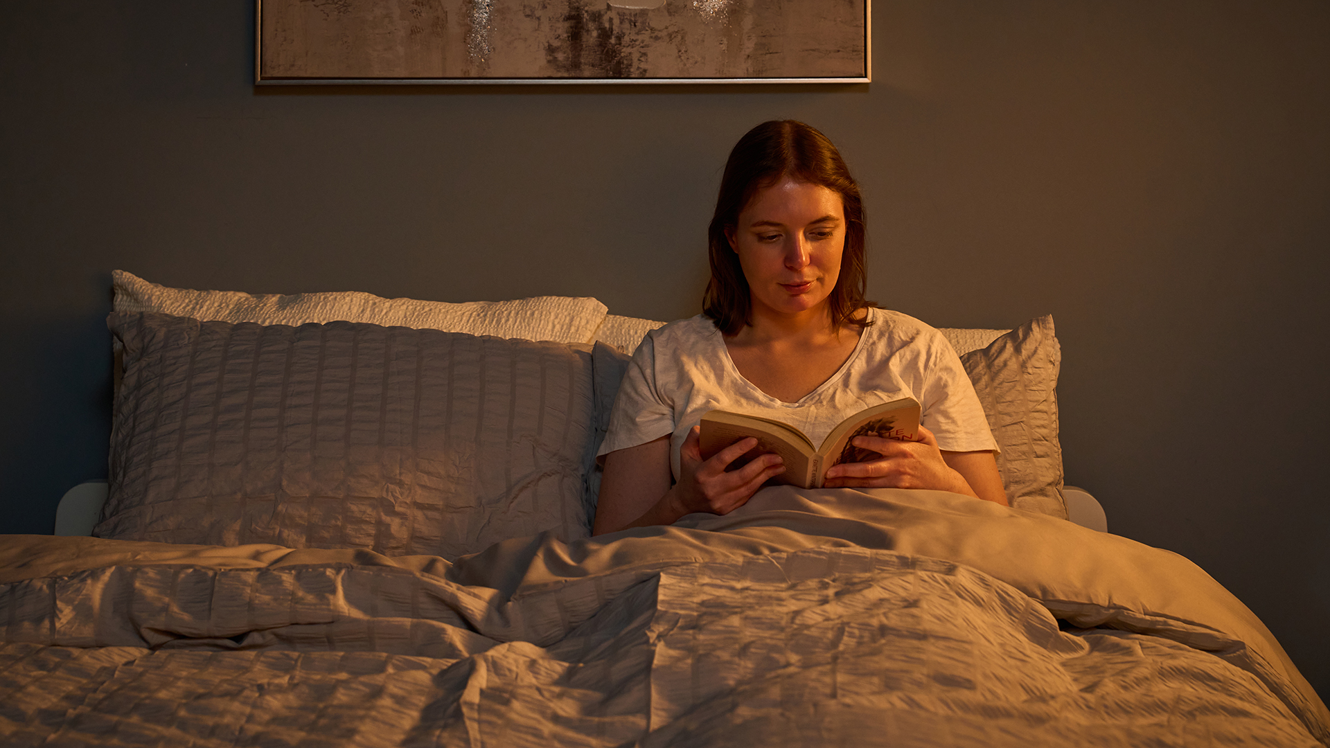 A woman sits in bed reading a book in dim light