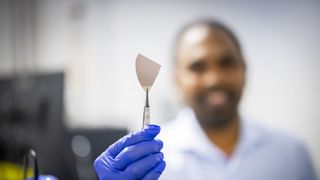 A photo of a material science researcher holding a piece of AlScN-based flash memory in tweezers, with the person out of focus in the background