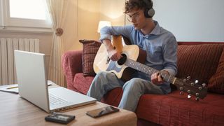 Man wearing headphones and holding an acoustic guitar takes a lesson via his laptop