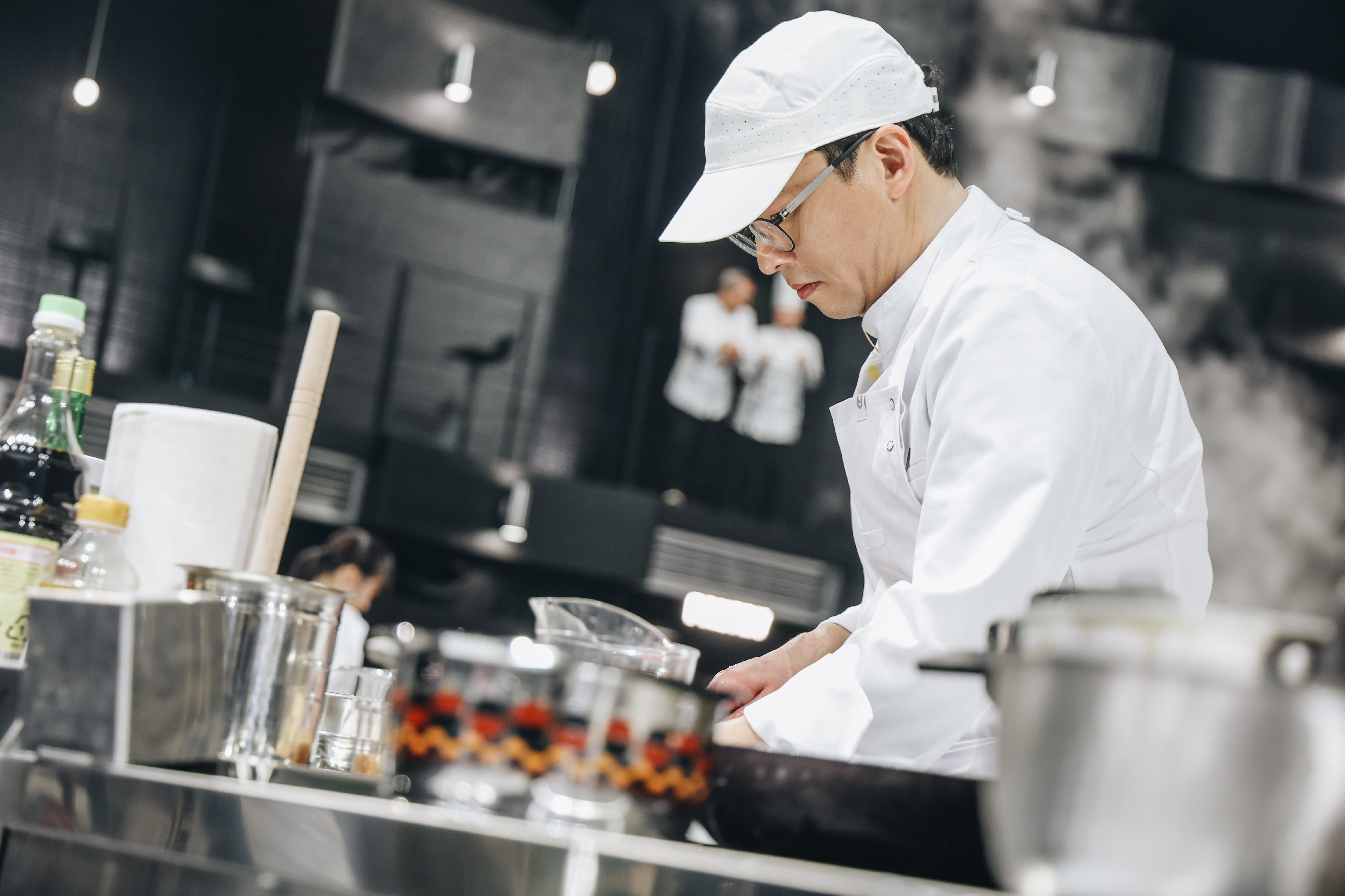 A chef (Choi Kang-rok) wears a cap and glasses as he cooks at a station, in 'Culinary Class Wars' season 2.