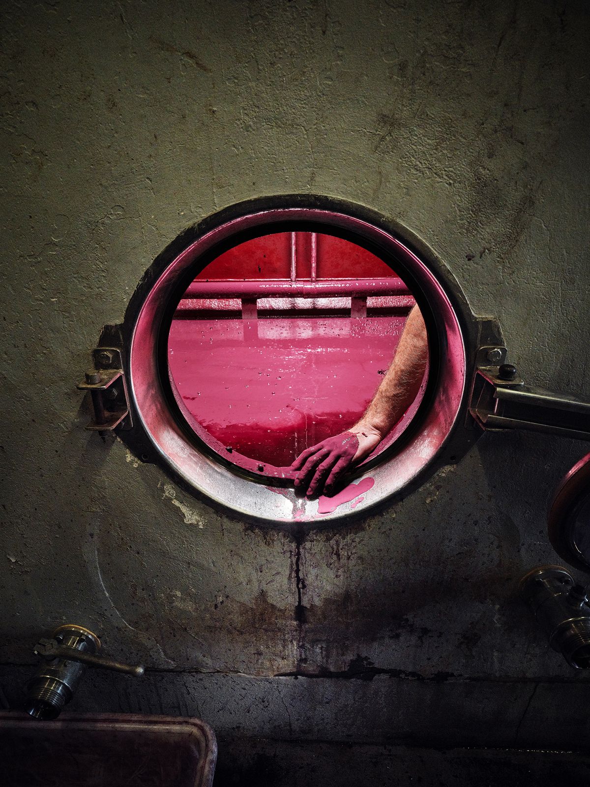 A hand reaches through a round opening, stained with deep purple liquid, inside a wine fermentation tank
