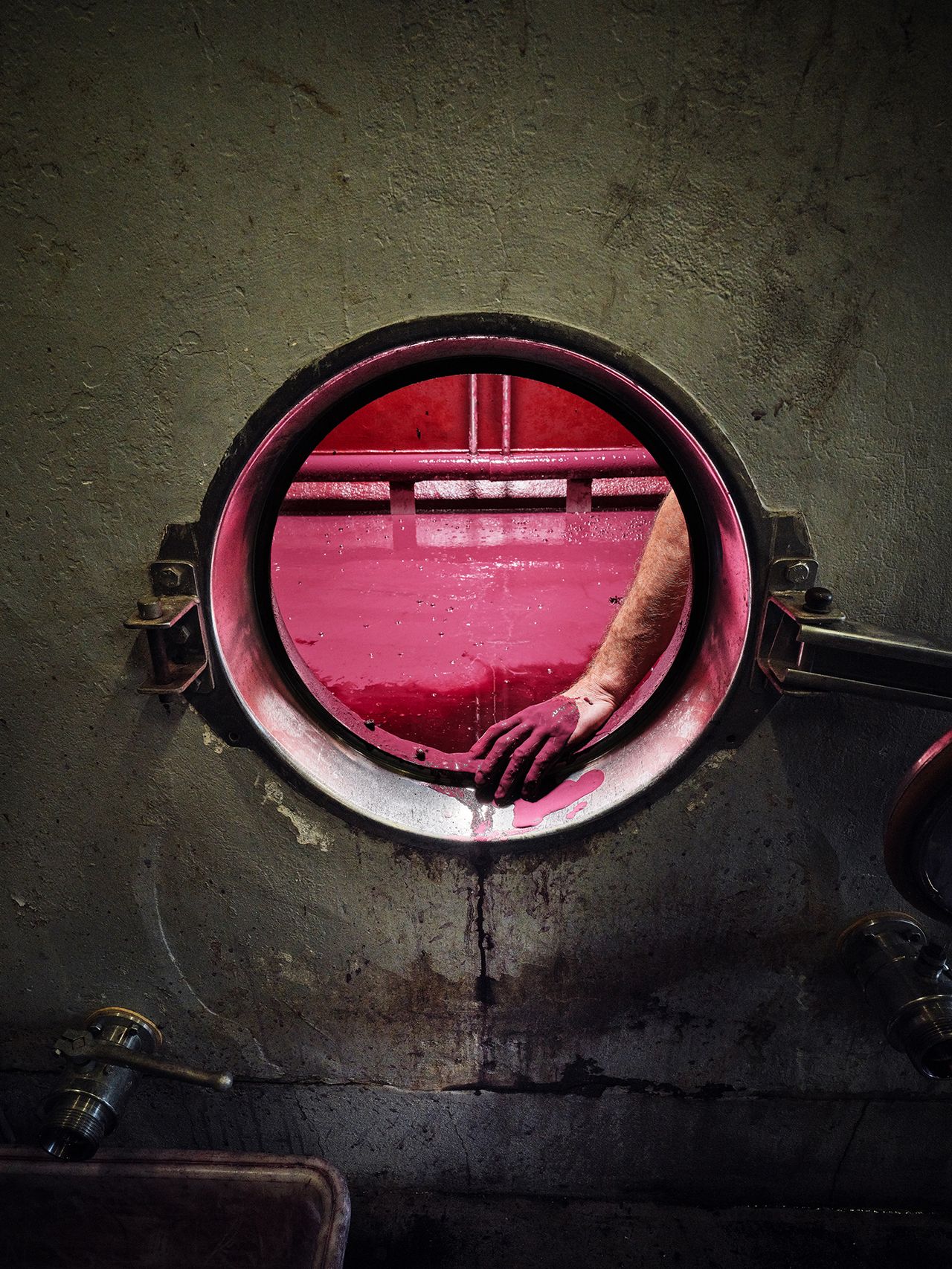 A hand reaches through a round opening, stained with deep purple liquid, inside a wine fermentation tank