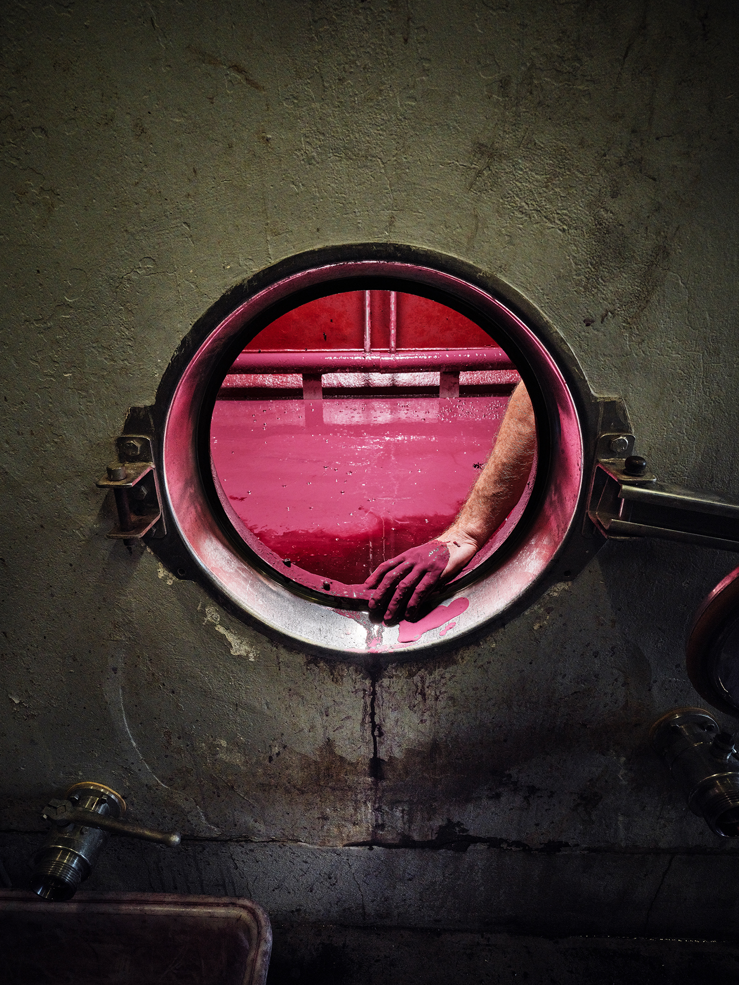 A hand reaches through a round opening, stained with deep purple liquid, inside a wine fermentation tank