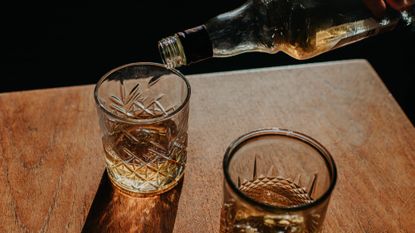 3/4 shot of two tumblers being filled with whiskey on a wood tabletop