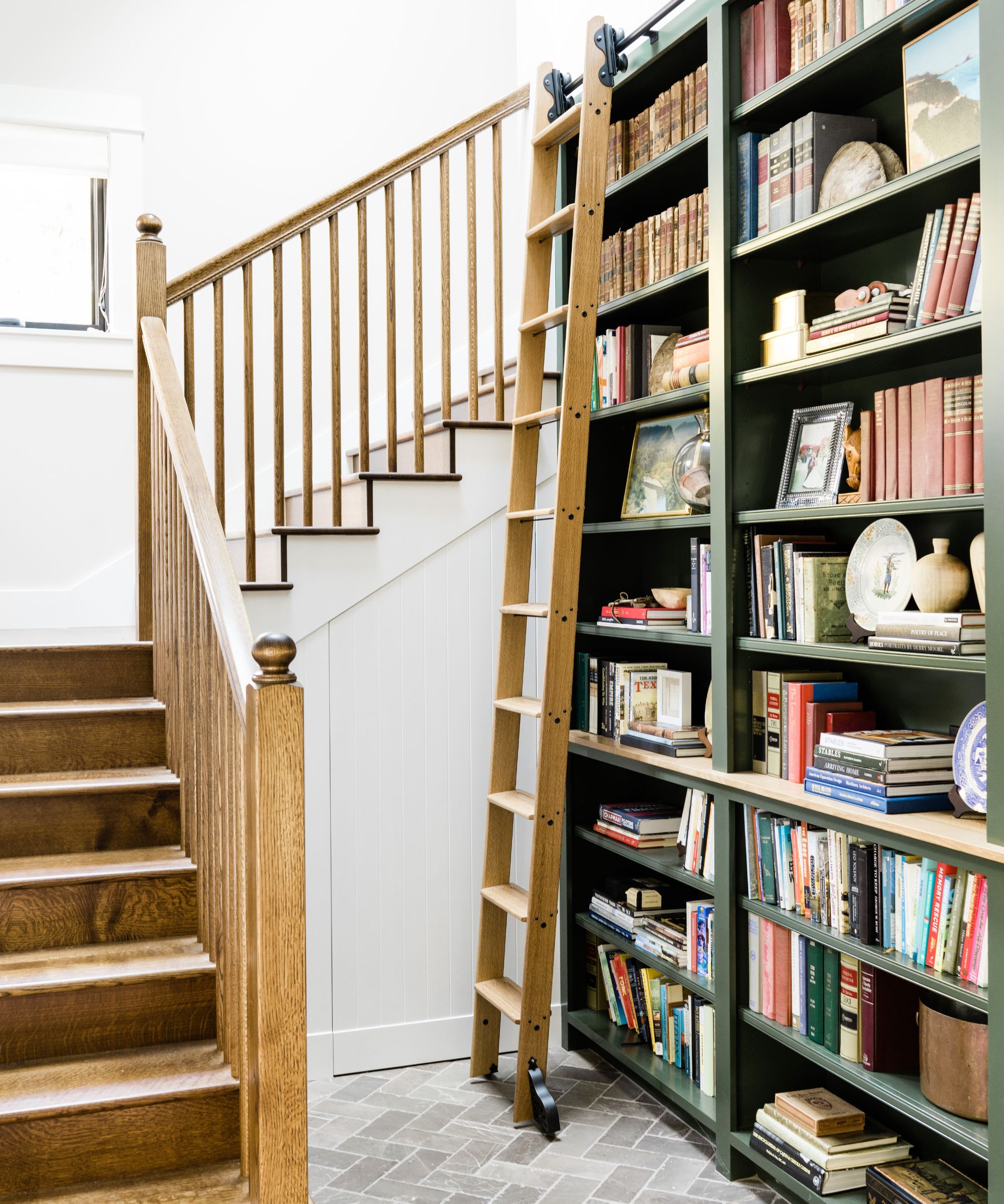 A hallway with a wooden staircase, white walls, and a dark green tall bookcase with a wooden ladder.