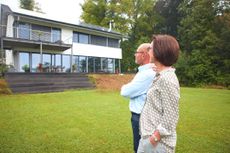 An older couple stands in their backyard, looking at their modern home. There are steps leading up to the porch with no bannister, indicating the home may not be ideal for aging in place.