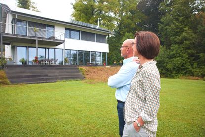 An older couple stands in their backyard, looking at their modern home. There are steps leading up to the porch with no bannister, indicating the home may not be ideal for aging in place.