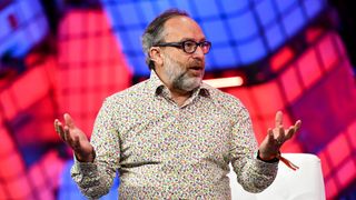 Jimmy wales sat down at a tech conference, speaking on a panel with a red and blue neon background behind him