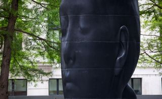 A large black sculpture of a human head with its eyes closed, positioned in the midst of tall trees and building. photographed during the day