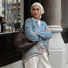 Woman wears blue cardigan, blue scarf, cream trousers and carries brown woven leather Dragon Diffusion Egola bag