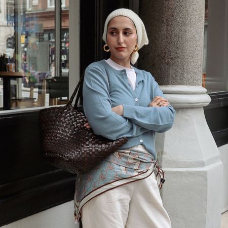 Woman wears blue cardigan, blue scarf, cream trousers and carries brown woven leather Dragon Diffusion Egola bag