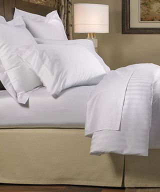 White bedding on a beige bed frame; in the background, an ornate table lamp.