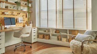 Bright, cosy home office with wooden floors, a large desk, shelves, woven baskets, and a plush armchair beside wide windows with white blinds.