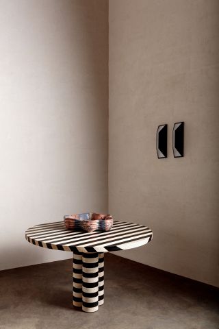 Round striped (black and white) dining table with 3 grouped cynlinder shaped legs (in the center) with a hand-molded colourful bowl placed on the table. Photographed in the corner of a room with white wall and brown floor
