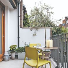 A small balcony with a round bistro table, two chartreuse chairs and potted plants, as well as a botted olive tree