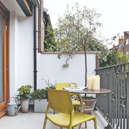 A small balcony with a round bistro table, two chartreuse chairs and potted plants, as well as a botted olive tree