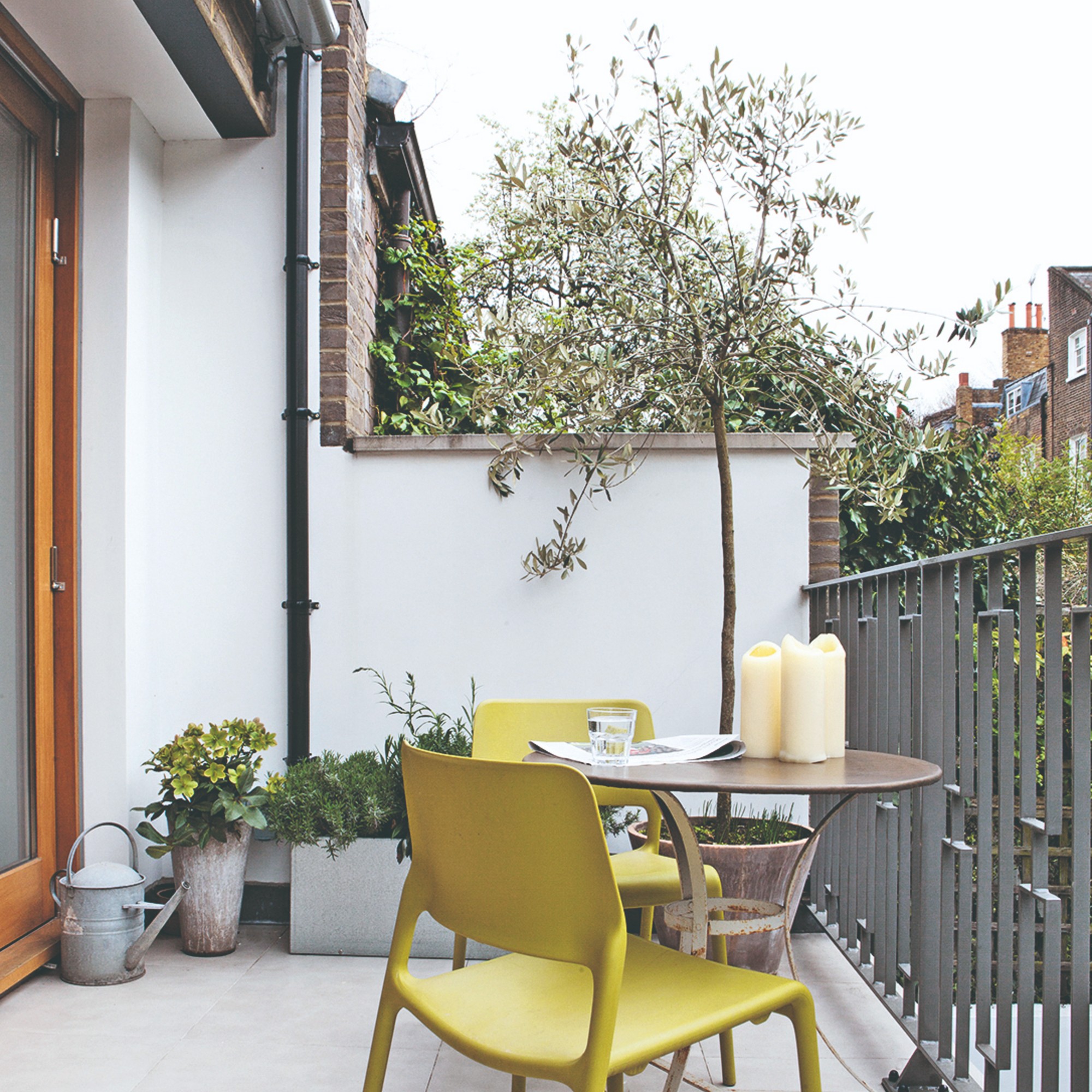 A small balcony with a round bistro table, two chartreuse chairs and potted plants, as well as a botted olive tree