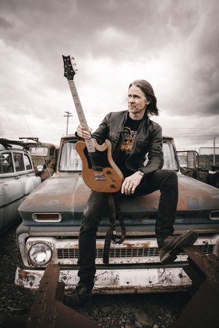 Myles Kennedy with guitar, sitting on a wrecked car