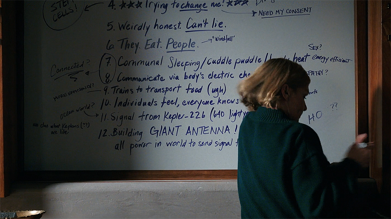 Carol standing in front of a whiteboard with information about The Others written on it in Pluribus episode 8