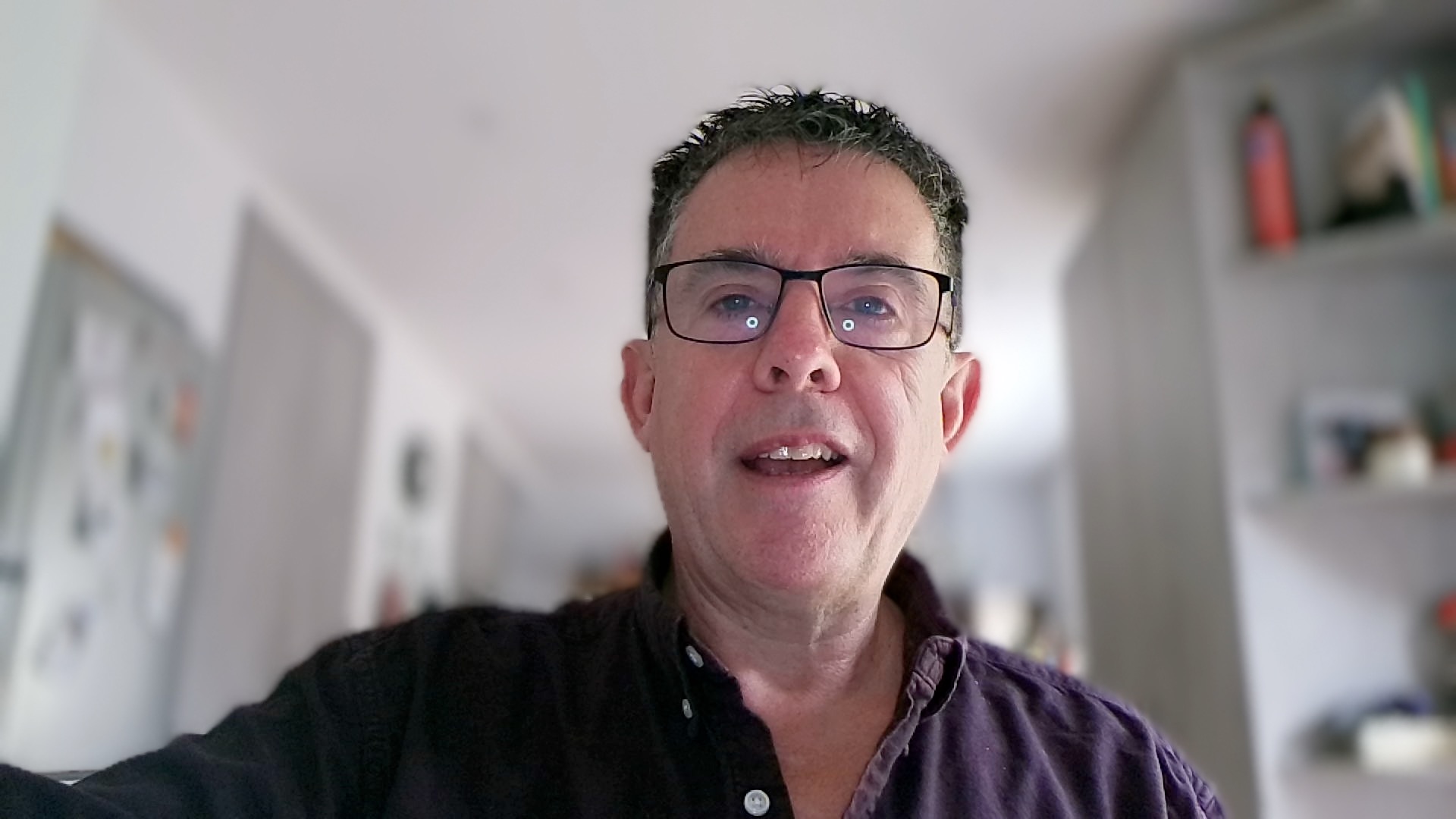 Webcam footage of a smiling man wearing glasses against a blurred indoor background.