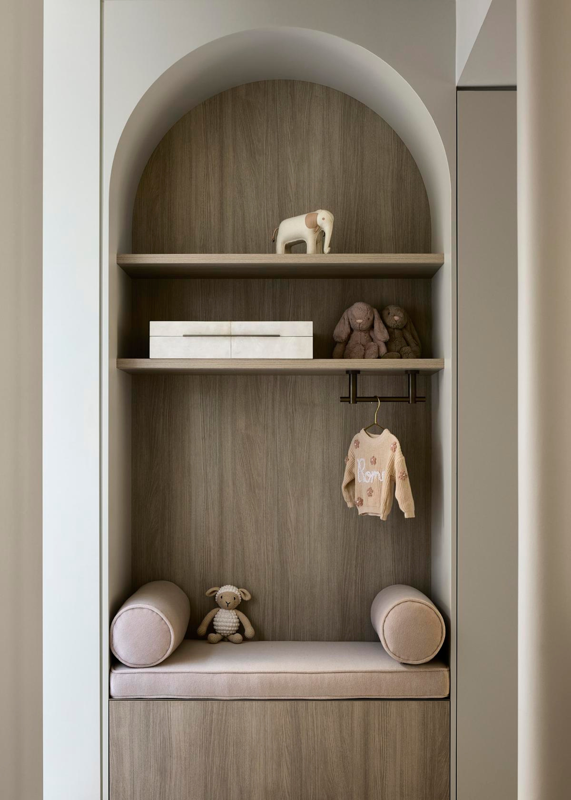 A muted kid's room with a domed nook featuring shelving for toys and a bench with bolster pillows, plus a hanging attachment for outfits