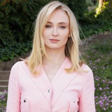 Sophie Turner smiles at the camera wearing a pink jacket