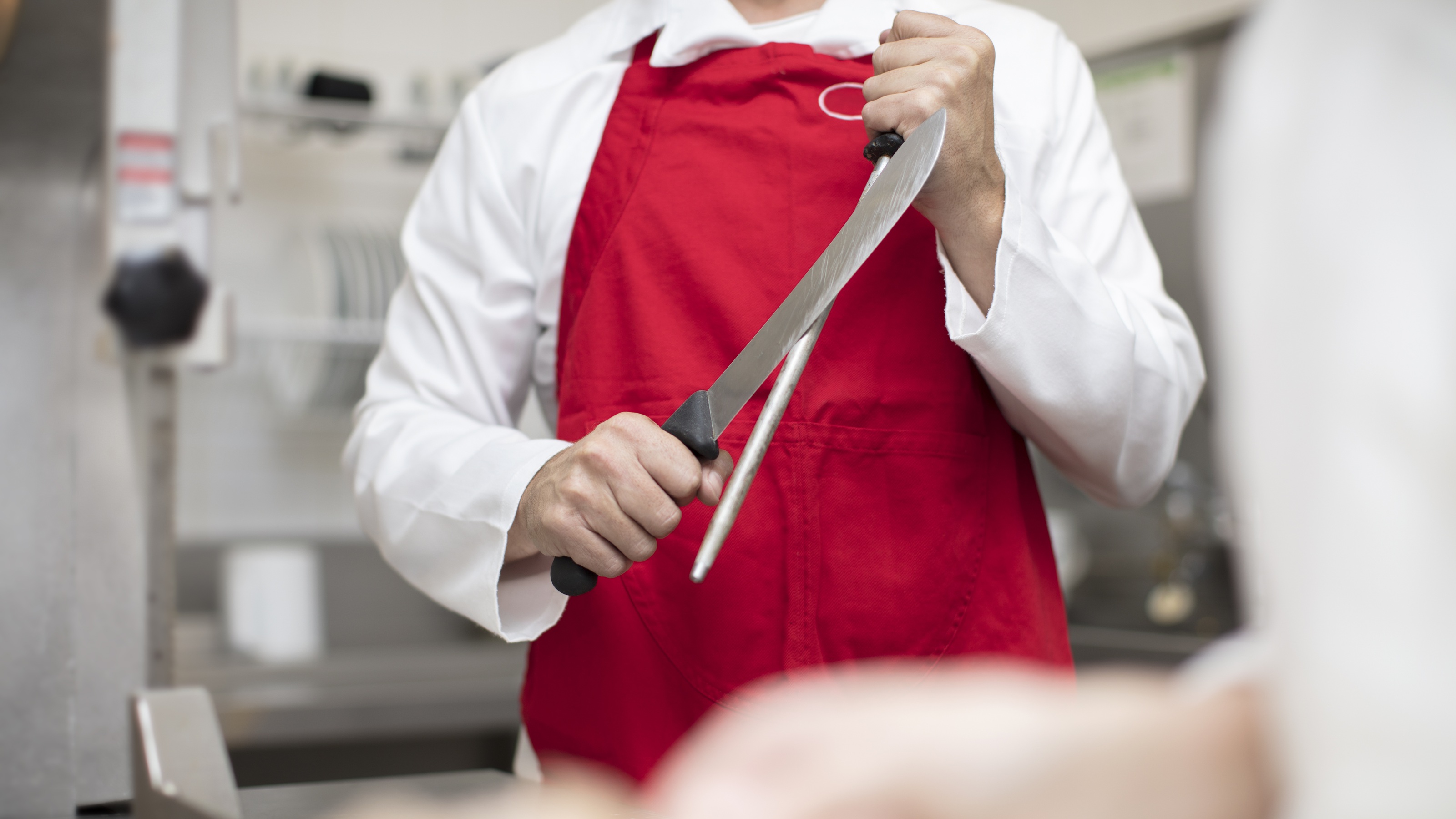 A chef in a red apron sharpens a knife in his commercial kitchen.