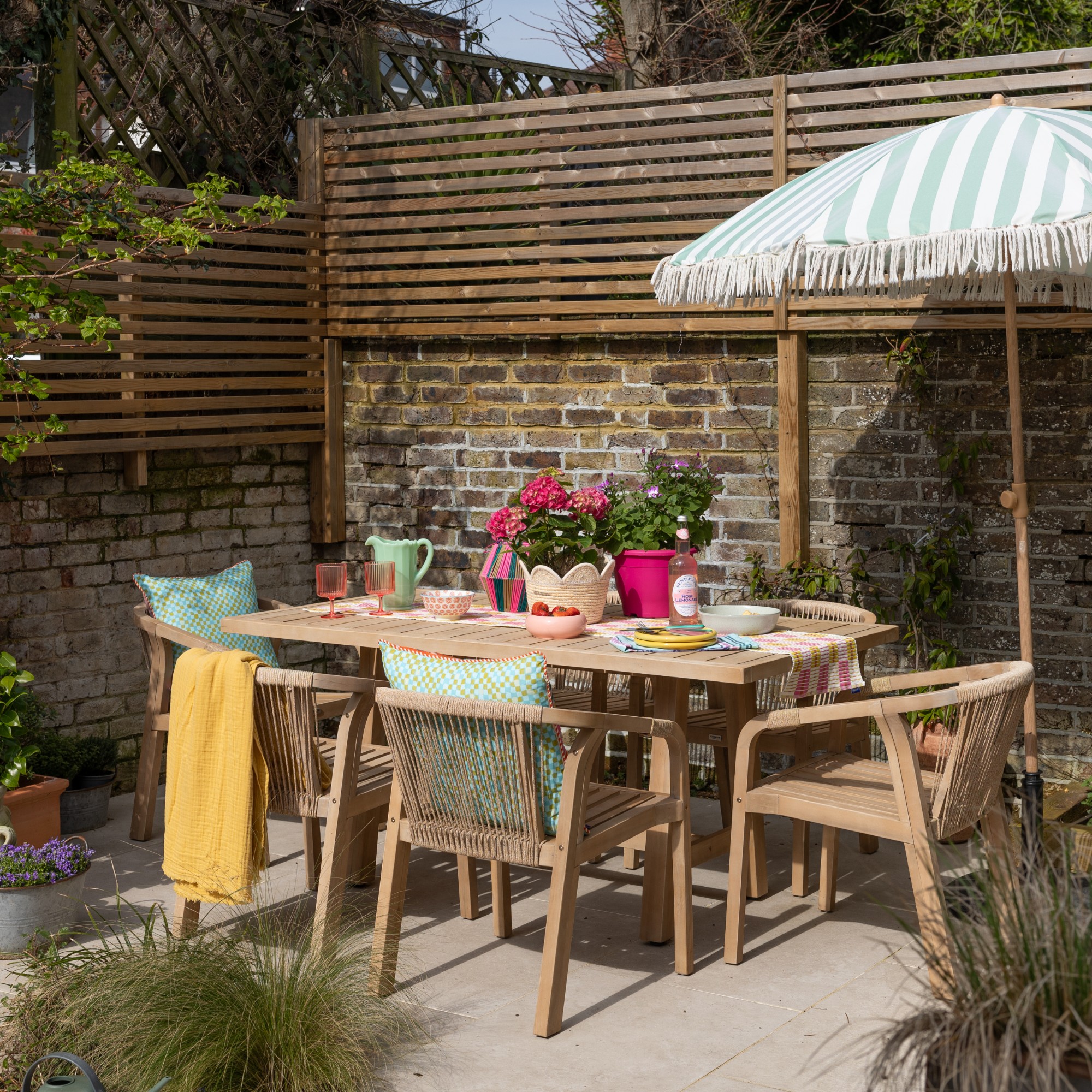 A garden with a wooden outdoor dining set with rope detailing and a striped, fringe-trimmed parasol next to it