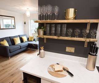 An open plan kitchen with wooden cutting board on a white stone island, leading to living room with dark blue sofa and mustard yellow cushions