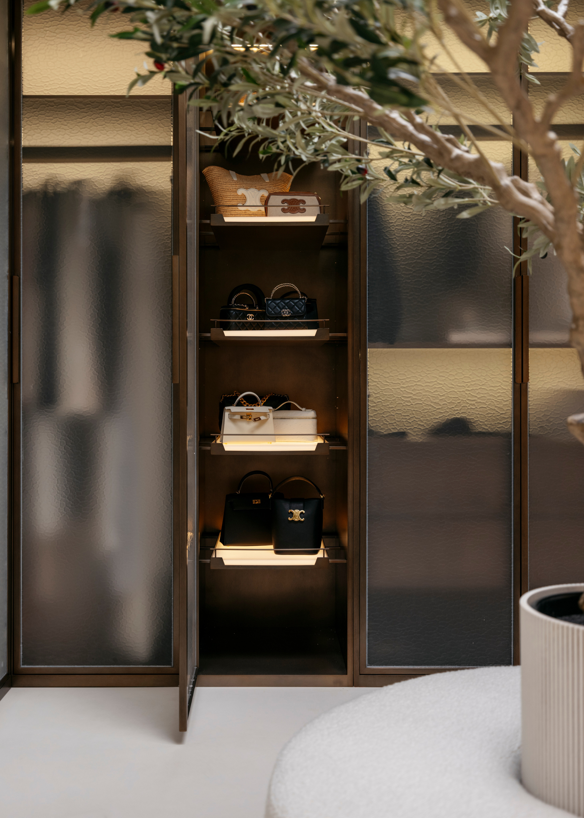 A minimalist wood wardrobe with frosted glass revealing vertically stored handbags and a glimpse of bench seating in front of a faux olive tree