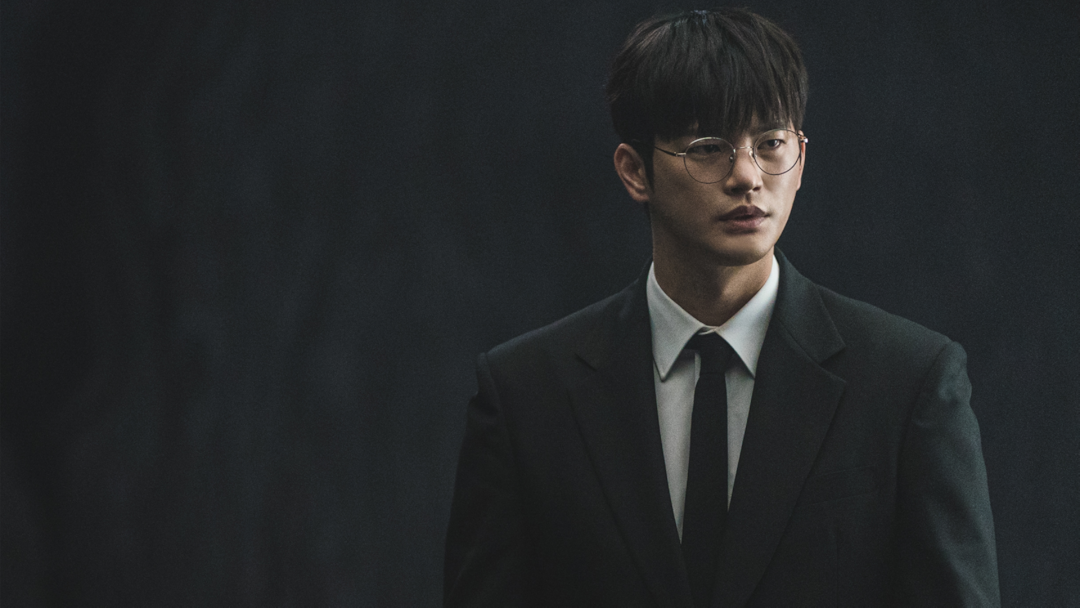 Seo In-guk in the Prime Video k-drama 'Death's Game' wearing circular glasses and a suit and tie against a black backdrop