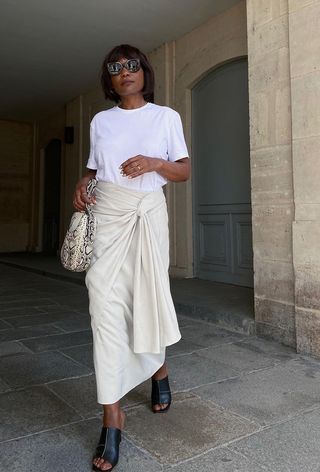 A snakeskin purse is shown in a street-style photo of a woman standing outside wearing black sunglasses, a white t-shirt, a tan wrap skirt, black mules, and a python bucket bag