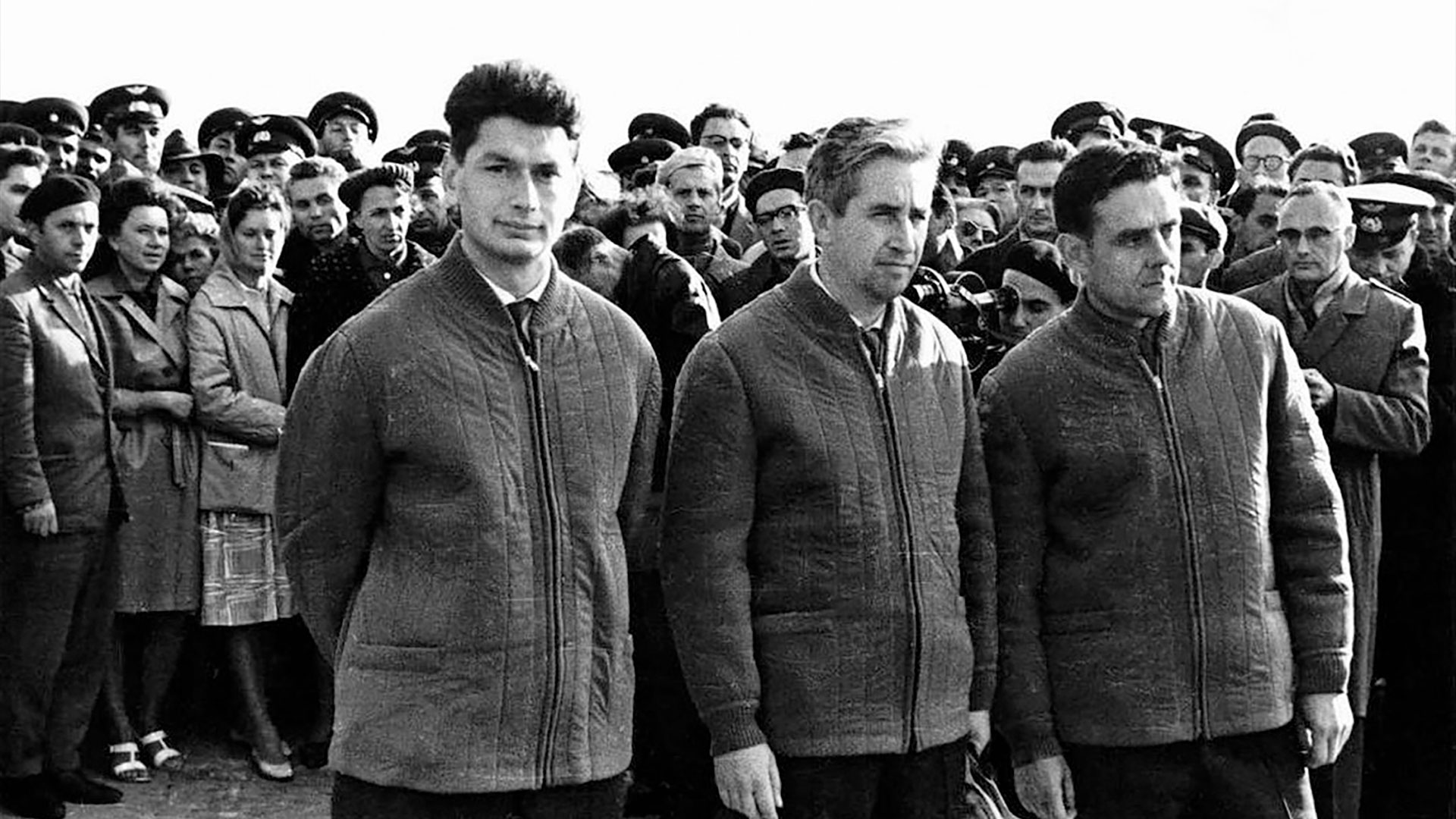 three men in jackets pose together in front of a large crowd of people