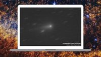 The ghostly light of a distant comet is pictured on the screen of a laptop as it travels across a distant starscape. The words 'Interstellar Comet 3I/ATLAS online observing session are visible to the lower right of the screen. The laptop is pictured against a deep space background composed of blue white stars and puffs of interstellar dust and gas.