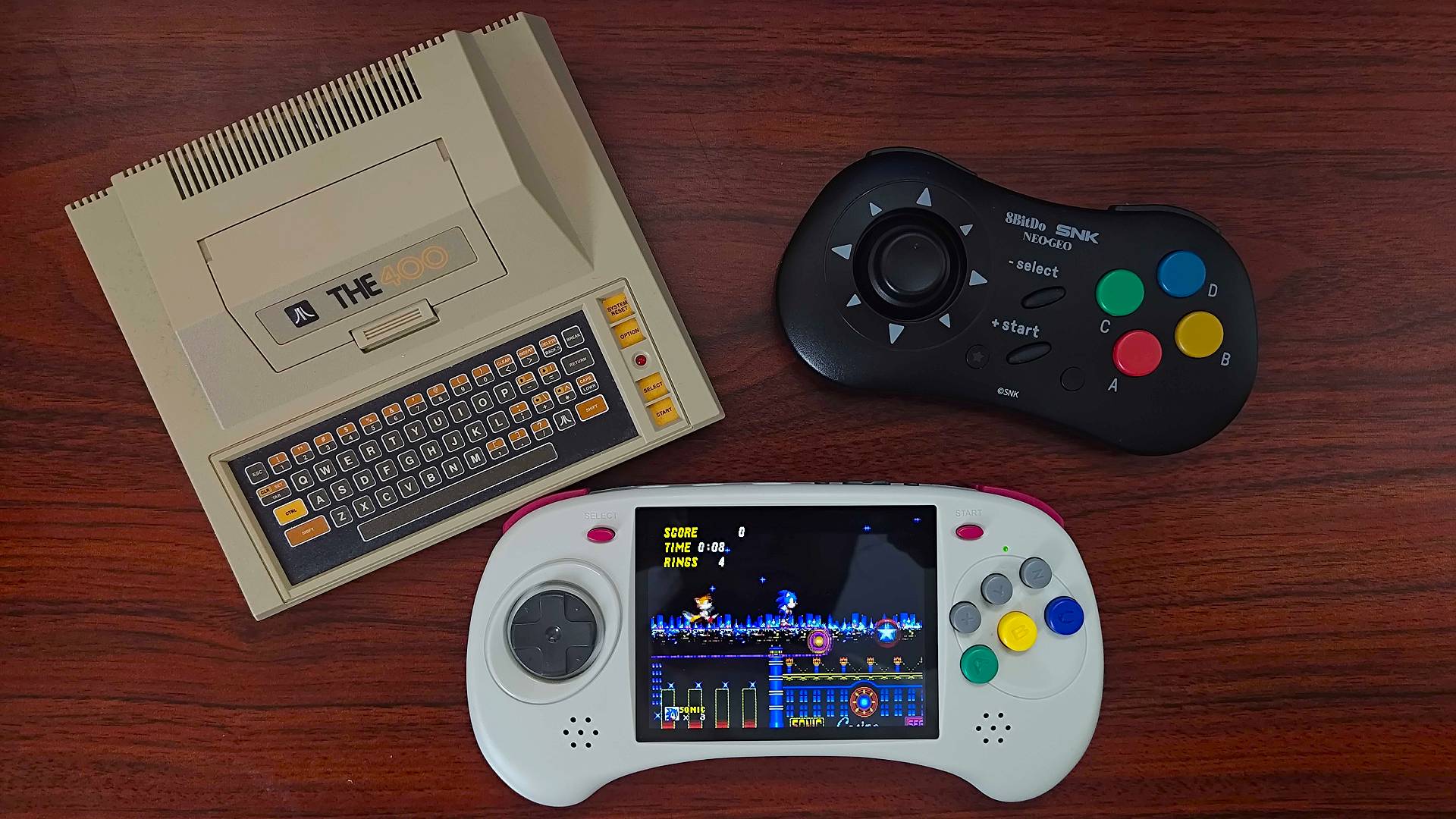 Atari 400 Mini, Anbernic RG ARC-D, and 8bitdo Neo Geo controller on woodgrain desk.