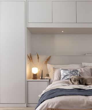 Neutral hued integrated built-in storage around bed, with bedside lamp and dried grasses in vase.