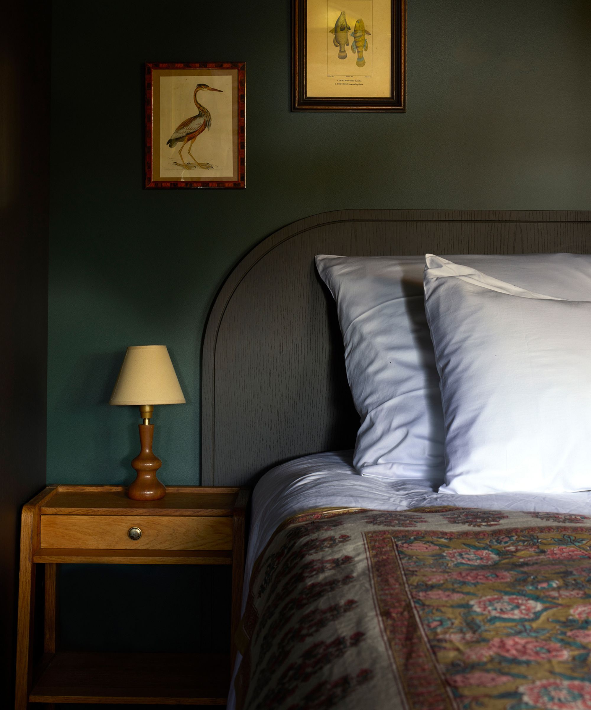 a dark moody green painted bedroom with a dark headboard, wooden nightstand and vintage artwork