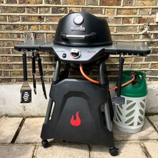 a gas barbecue againts a brick wall with a large gas canister beside
