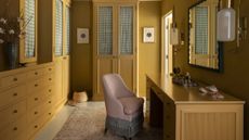 A mustard-yellow walk-in wardrobe, designed by Nickey Kehoe, with built-in cabinetry along the walls, and a pink upholstered chair in front of a large desk.