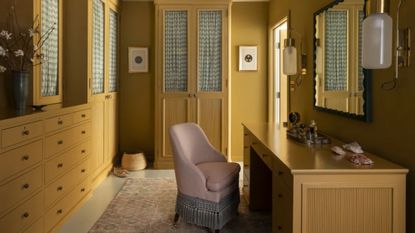 A mustard-yellow walk-in wardrobe, designed by Nickey Kehoe, with built-in cabinetry along the walls, and a pink upholstered chair in front of a large desk.