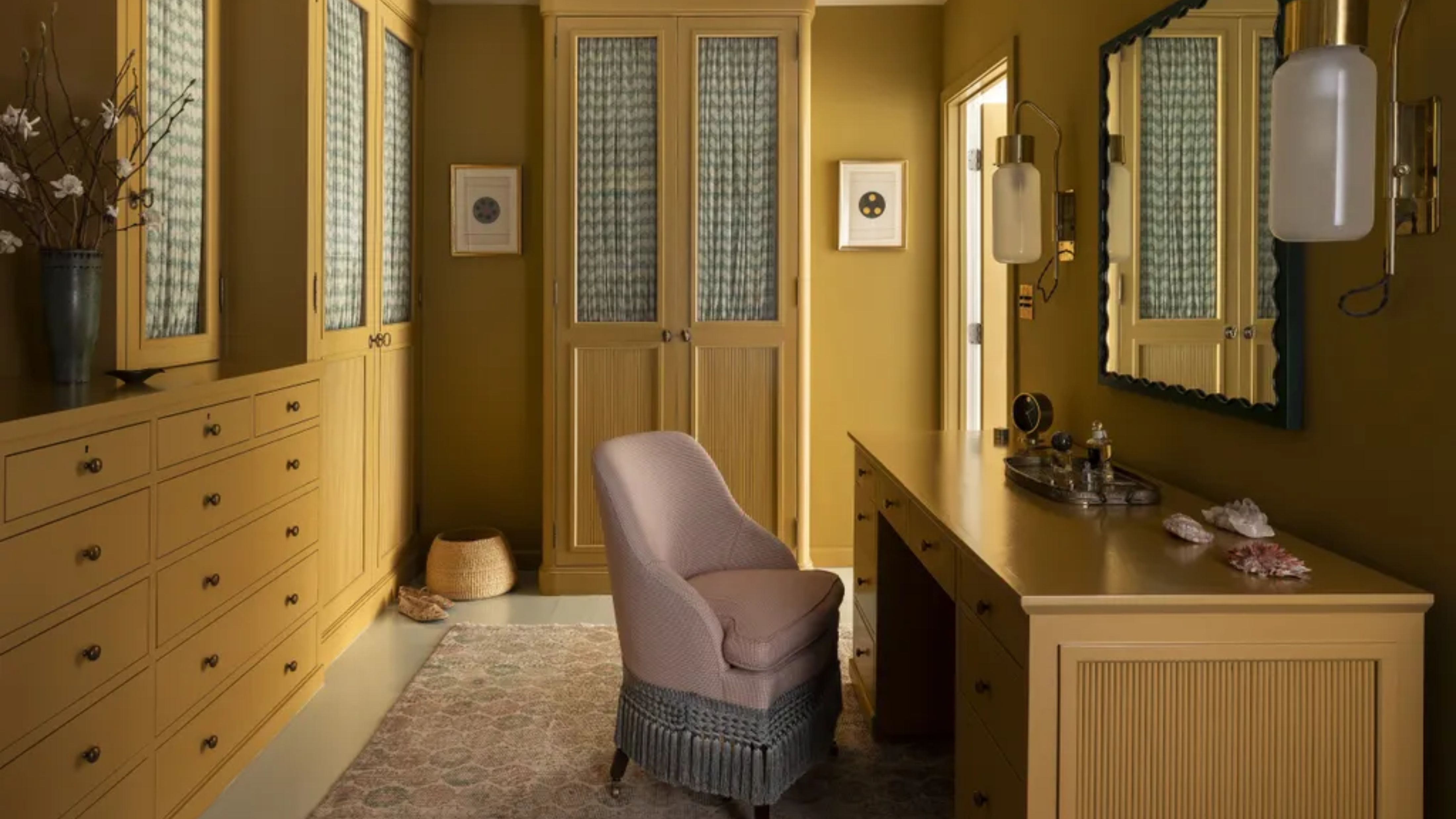 A mustard-yellow walk-in wardrobe, designed by Nickey Kehoe, with built-in cabinetry along the walls, and a pink upholstered chair in front of a large desk.