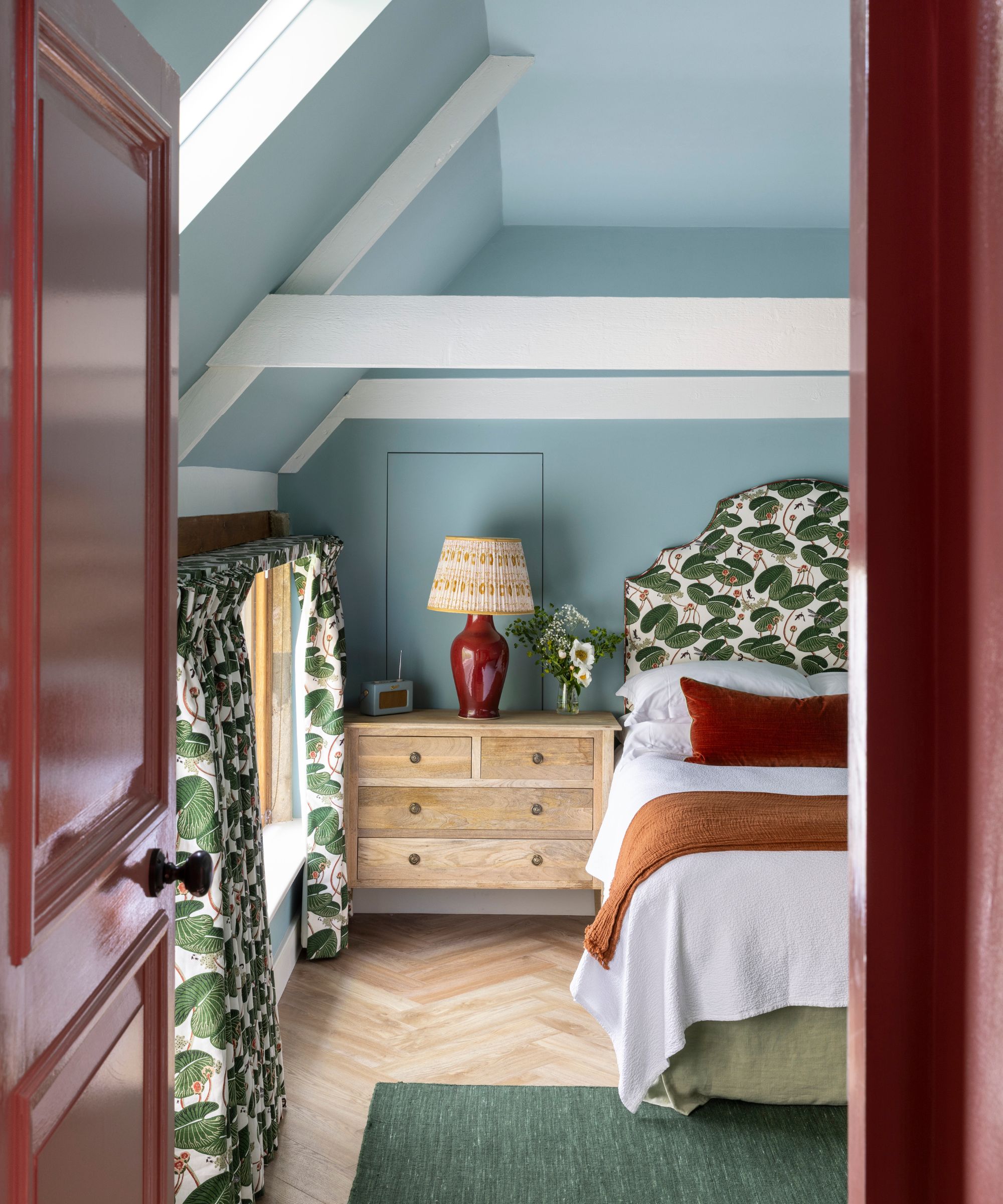A bedroom with a sloped ceiling and light blue walls, a patterned headboard, and red and brown textiles.