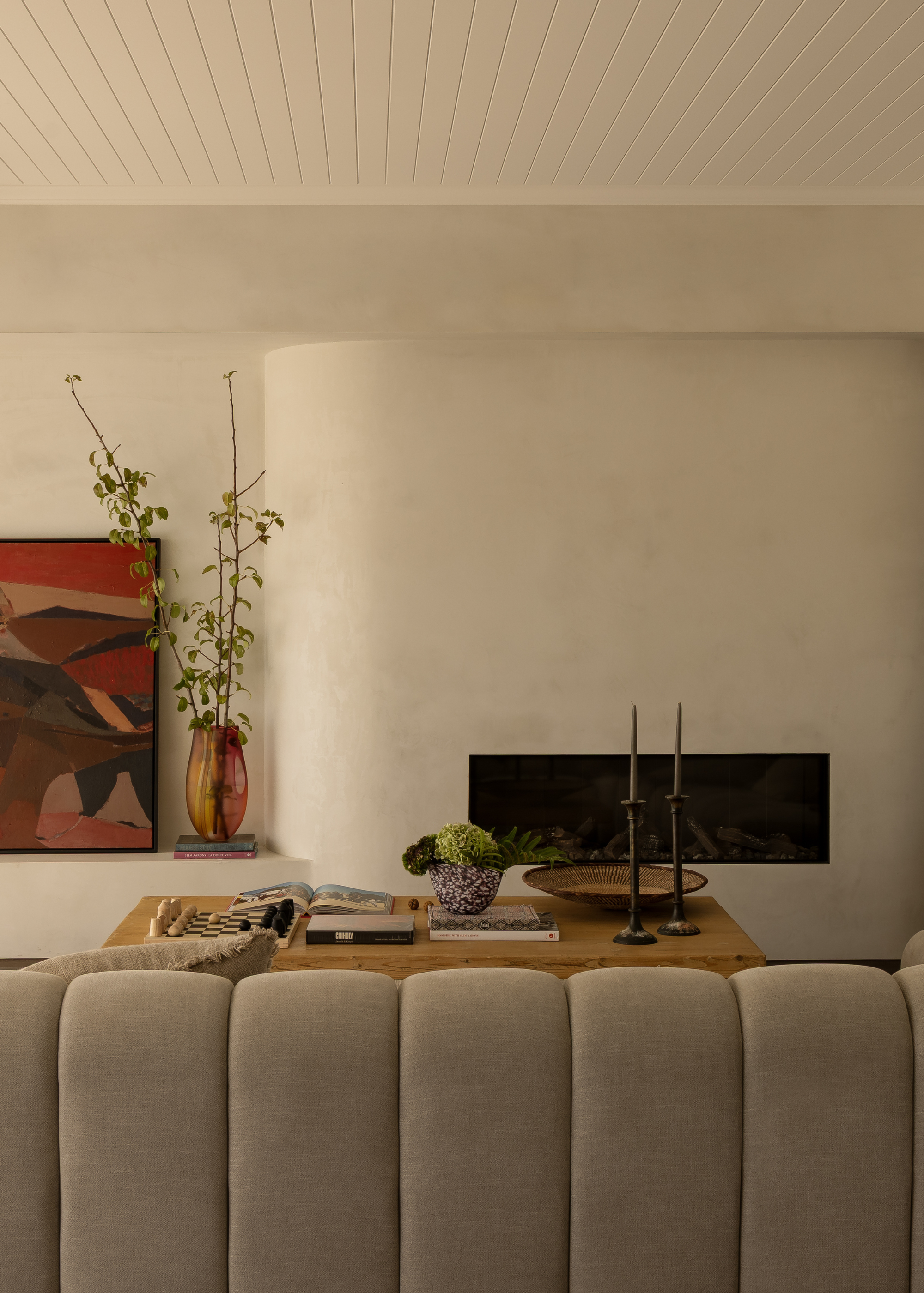 A white living room with a panelled ceiling, an in-built fireplace, a wood coffee table with taper candles, a vase of hydrangeas, a couple books, a chess set, and a bowl