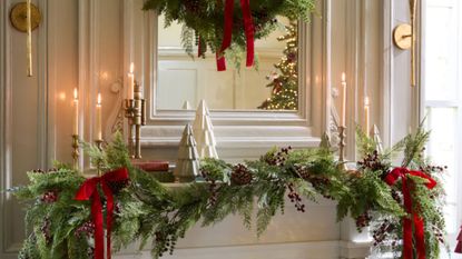 Festive fireplace mantel featuring evergreen garland, lit taper candles, ceramic Christmas trees, and red bows.