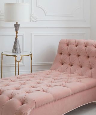 A dusky pink chaise lounge with a button tufted pattern, a gold and glass table next to it with a gray hourglass lamp with a white lampshade on it next to it