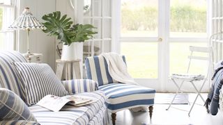 White painted living room with coastal style blue striped small soaf and armchair with French doors looking out to a garden