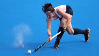 Amy Costello of Team Great Britain, in white vest, navy blue shorts and black socks, passes the ball along the blue astro hockey pitch at the 2024 Paris Olympic Games.