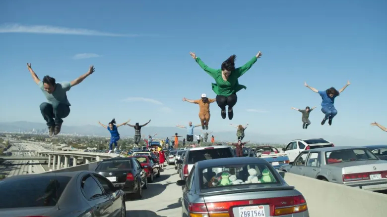 La La Land still. Rows of cars at a standstill on a motorway under a blue sky. People are mid-air with their arms up and are jumping on the roof of each car.