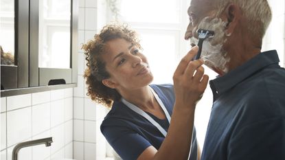 An older man gets help shaving from a health care aide.